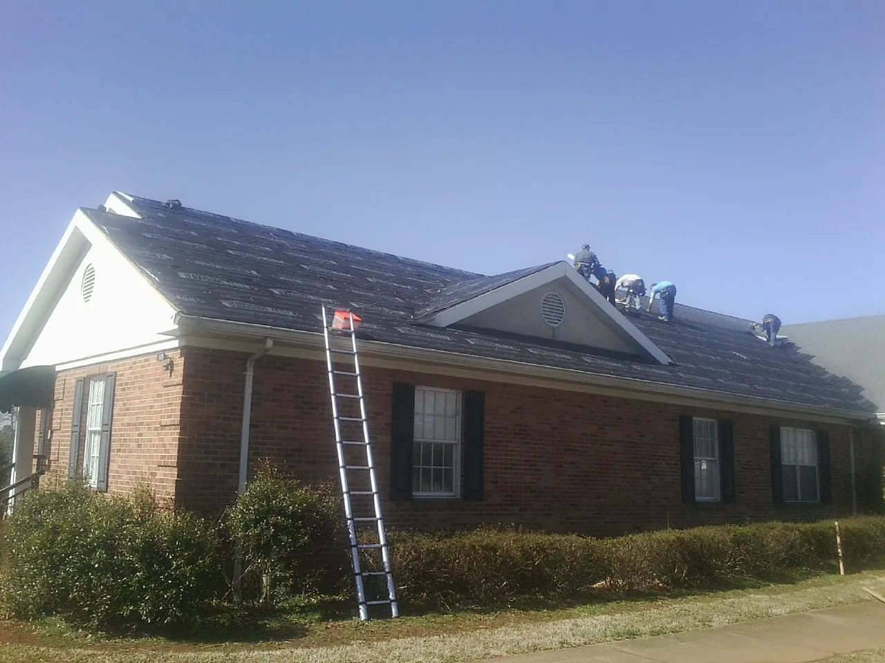 Crew tearing shingles off a brick ranch roof with a ladder set — Emergency Roof Repair work by our crew in Stitzer