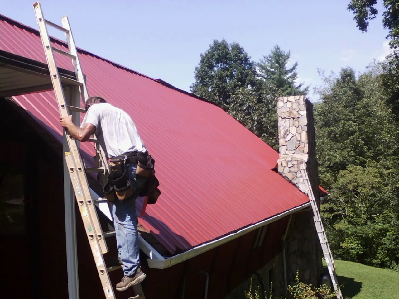 Roofer on a ladder beside a red metal roof and stone chimney — Soffit & Fascia Repair by Roofing Brothers in Stitzer